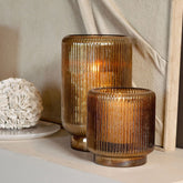 Set Van 3 Shalea Windlichten Amber Ribbelglas van PTMD, twee windlichten met brandende kaarsen op een sokkel tegen een lichte muur